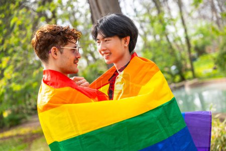 Photo for Multi-ethnic gay couple embraced in a park wrapped in rainbow flag - Royalty Free Image