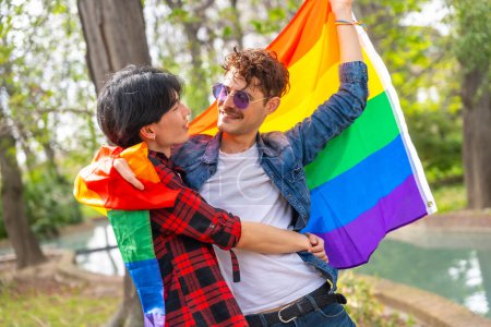 Glückliches und romantisches multiethnisches schwules Paar, das sich in Regenbogenflagge in einem Park umarmt