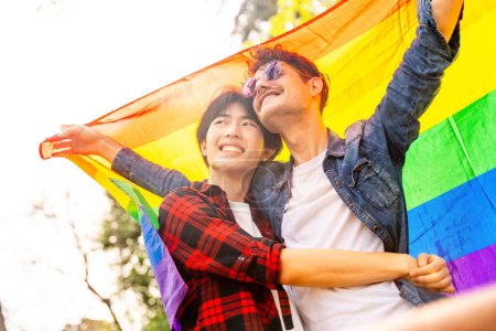 Photo for Low angle view of a multi-ethnic gay couple celebrating diversity raising lgbt flag in a park - Royalty Free Image