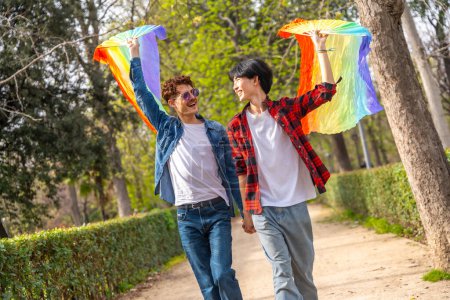 Photo for Happy multi-ethnic young gay couple raising LGBT rainbow colored fans in a park - Royalty Free Image