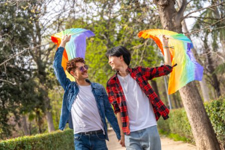Photo for Happy multiracial gay couple walking holding hands and waving LGBT rainbow hand fans in a park - Royalty Free Image