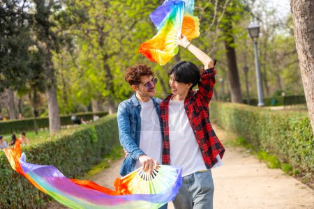 Photo for Multiracial gay couple waving lgbt rainbow fans while walking together along a public park - Royalty Free Image