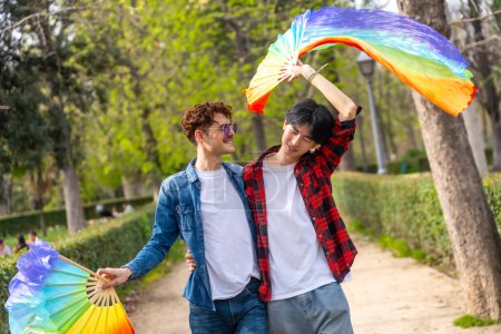Photo for Multiracial gay couple celebrating love waving lgbt rainbow fans in a park - Royalty Free Image