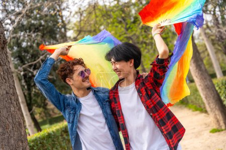 Photo for Multiracial gay couple raising lgbt rainbow hand fan smiling walking in a park - Royalty Free Image