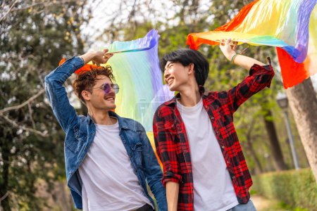 Photo for Gay couple holding hands strolling happily in a park raising lgbt rainbow hand fans - Royalty Free Image