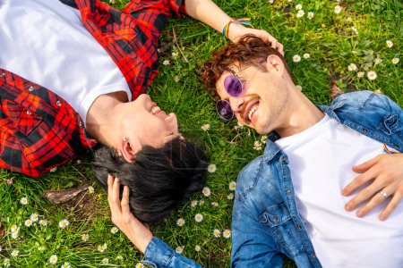 Foto von oben von einem multiethnischen homosexuellen Paar, das gemeinsam auf dem Rasen eines Parks liegt und einander ansieht und lächelt
