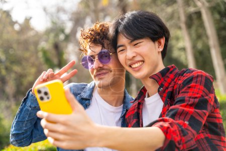 Photo for Close-up of a multiracial gay couple gesturing success while taking selfie in a park - Royalty Free Image