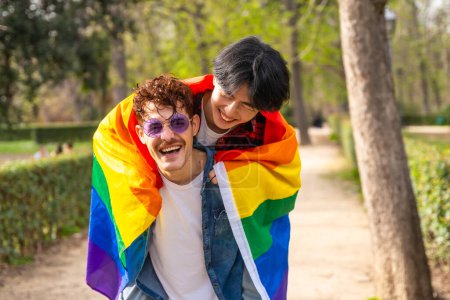 Photo for Multi-ethnic gay men wrapping in lgbt flag piggybacking in a park - Royalty Free Image