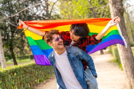 Multi-ethnische schwule Paar huckepack in einem Park eine lgbt Regenbogenfahne hissen
