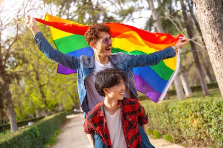 Photo for Multiracial gay couple waving lgbt flag piggybacking celebrating diversity in an urban park - Royalty Free Image