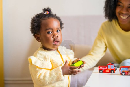 Téléchargez les photos : Petite fille africaine mignonne jouant à des jouets avec sa mère dans le salon à la maison - en image libre de droit
