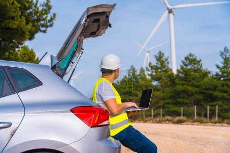 Trabajador con ropa protectora usando laptop trabajando en un parque de energía eólica sentado en la bota del auto