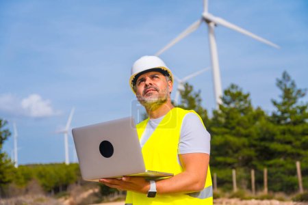 Male worker with protective clothes using laptop standing in a wind turbine field in a sunny day