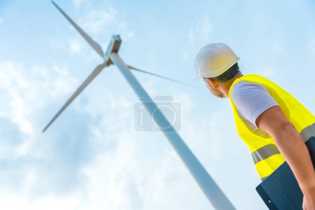 Low angle view photo of a male caucasian adult worker in protective gear looking up at a wind turbine