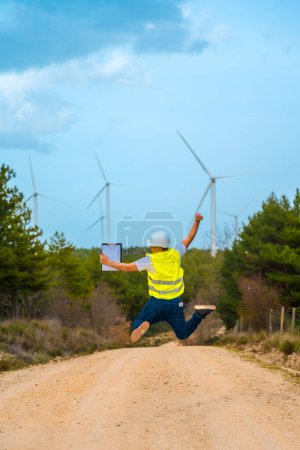 Vue arrière verticale d'un ingénieur adulte caucasien célébrant un saut en l'air à côté d'éoliennes dans un parc