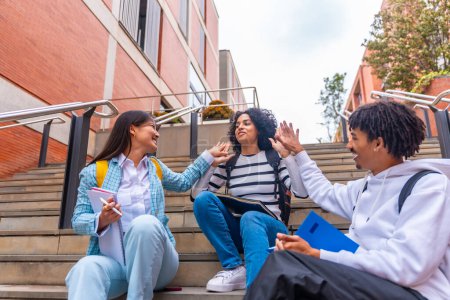 Téléchargez les photos : Multi-ethnique trois étudiants de haute-cinq tout en étant assis sur certains escaliers sur le campus universitaire - en image libre de droit