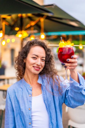 Vertical photo of a woman toasting at camera with a cocktail sitting on a bar terrace