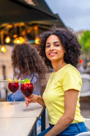 Vertical photo of a latin woman smiling at camera while enjoying cocktails with friends in an outdoor bar in summer