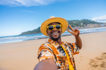 Afrikanischer Mann mit Sonnenhut, Sonnenbrille und bunten Klamotten gestikuliert Sieg beim Selfie am Strand