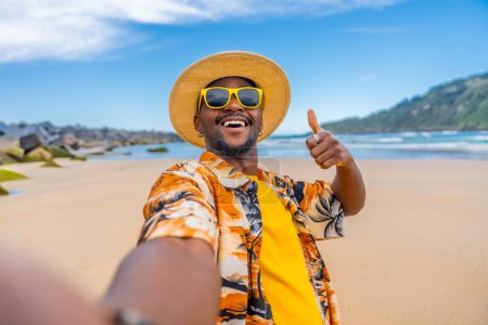 Cool man with summer clothes gesturing with thumb up taking a selfie on the beach
