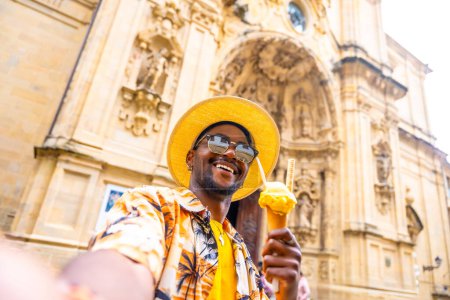 African male tourist in sun hat and colorful clothes eating an ice cream and taking selfie