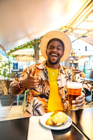 Vertical photo of a happy young male african american tourist enjoying beer and sandwich on a terrace