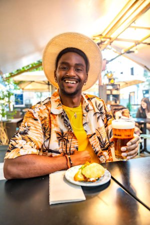 Vertical portrait of a young african american man in sun hat sitting on a sidewalk cafe enjoying beer and sandwich
