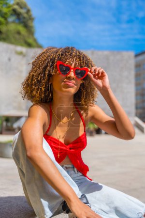 Vertical photo of a cute latin woman with curly hair using heart shape red sunglasses sitting in the city