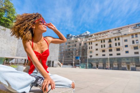 Horizontal photo with copy space of a beauty latin young fashionable woman sitting in an urban square enjoying sun wearing heart shape sunglasses
