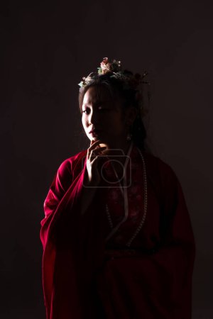 A woman in a red dress with a flowery headpiece is standing in a dark room. She is looking down and she is deep in thought