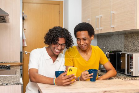 Multiracial gay couple sitting on the kitchen enjoying coffee cup and using phone