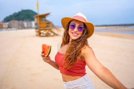 Eine Frau lächelt und hält eine Wassermelonenscheibe in der Hand. Sie trägt Strohhut und Sonnenbrille
