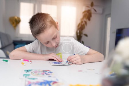 Téléchargez les photos : Un garçon de 4-7 ans s'assoit à la table et dessine avec des peintures multicolores. La créativité des enfants. Loisirs des enfants - en image libre de droit