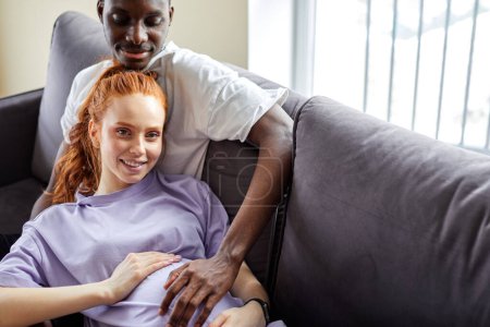 Téléchargez les photos : Lovely man stroking wifes pregnant belly, lying on sofa at home, relaxing. millennial black man and caucasian woman on comfortable couch, dream of bright happy future together in living room - en image libre de droit