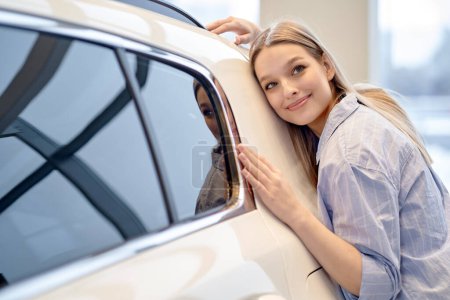 Téléchargez les photos : Pretty caucasian female hugging new car auto in dealership, enjoy the purchase or dream to get this one. attractive lady in casual wear in cars showroom, leaned on white expensive automobile - en image libre de droit