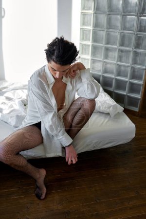 Téléchargez les photos : Somnolent jeune gay assis dans la chambre à coucher, regarde vers le bas, pleine longueur photo, mode - en image libre de droit
