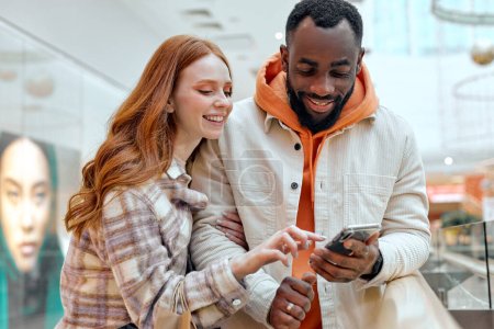 Photo for Young happy girl pointing to the screen showing her bearded boyfriend sales couple choosing goods, lifestyle.hobby interests red-haired girl showing something on smart phone to handsome boyfriend - Royalty Free Image