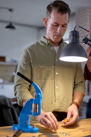 Téléchargez les photos : Tailleur décorant la ceinture faite à la main. homme utilise outil de coupe de métal, machine d'emboutissage pour l'extrusion de la peau. - en image libre de droit