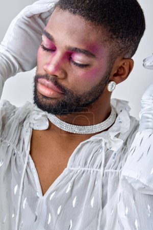 Photo for Pacified black man with make-up touching face head, in white gloves, elegant and gorgeous male in white shirt posing as professional model, looking feminine or womanlike, princess, cute - Royalty Free Image