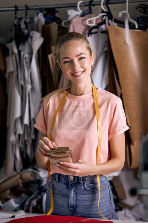Photo for Attractive caucasian dressmaker woman sews clothes at tailor office, engaged in tailoring, blonde female in casual wear posing at camera, holding fabric in hands. dressmaking industry - Royalty Free Image