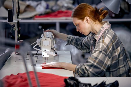 Photo for Seamstress lady in casual wear sit behind table using sewing machine, making modern clothes, young redhead caucasian woman at workshop, holding red fabric, enjoy tailoring, controlling equipment - Royalty Free Image