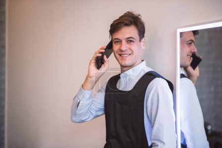 Foto de Retrato sonriente joven adulto caucásico masculino peluquero profesional usando teléfono inteligente para hablar con el cliente para la reserva de peluquería, Pequeña empresa en la ciudad. - Imagen libre de derechos