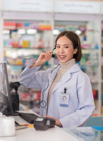 Photo for Smiling young asian pharmacist wearing headset to telemedicine online service with patient via computer at drug store. - Royalty Free Image