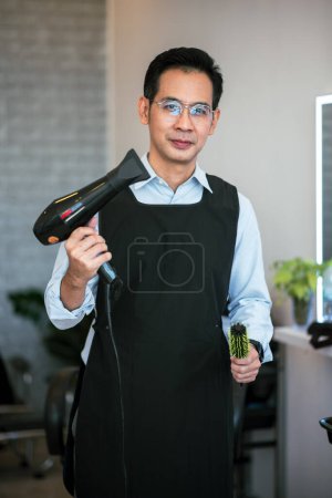 Téléchargez les photos : Portrait adulte asiatique homme coiffeur professionnel tenant sèche-cheveux et brosse à cheveux dans le salon de beauté - en image libre de droit