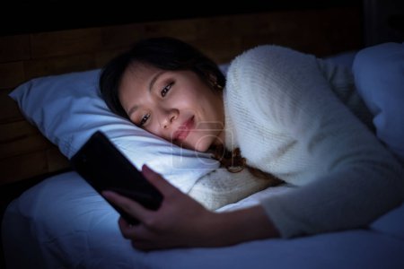 Foto de Mujer asiática sonriente acostada y usando smartphone para redes sociales en la cama, sin dormir por la noche, dormitorio con poca luz. - Imagen libre de derechos