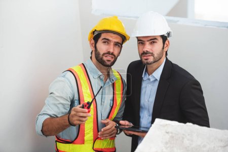 Foto de Hombres de trabajo en equipo en el sitio de construcción, dos ingenieros civiles en casco de seguridad casco duro usando tableta digital y plano de trabajo mientras están de pie en la fábrica. - Imagen libre de derechos