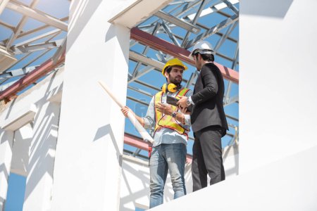Téléchargez les photos : Hommes de travail d'équipe sur le chantier, deux ingénieurs civils en casque de sécurité casque dur à l'aide d'une tablette numérique et un plan de travail tout en restant debout à l'usine. - en image libre de droit