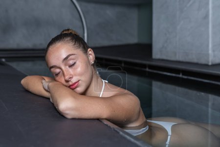 Photo for Young female in white bikini closing eyes and holding head on folded arms while chilling in transparent water of clean pool on spa resort - Royalty Free Image