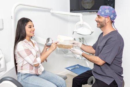 Photo for Side view of delighted ethnic teen girl with long dark hair in casual clothes with aligner box in hand taking prescription from hands of smiling male dentist during check up in clinic - Royalty Free Image