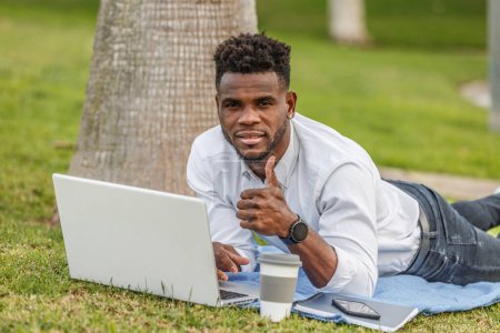 Photo for An African-American man is seen with his thumb up lying on the floor while using a laptop to work. - Royalty Free Image